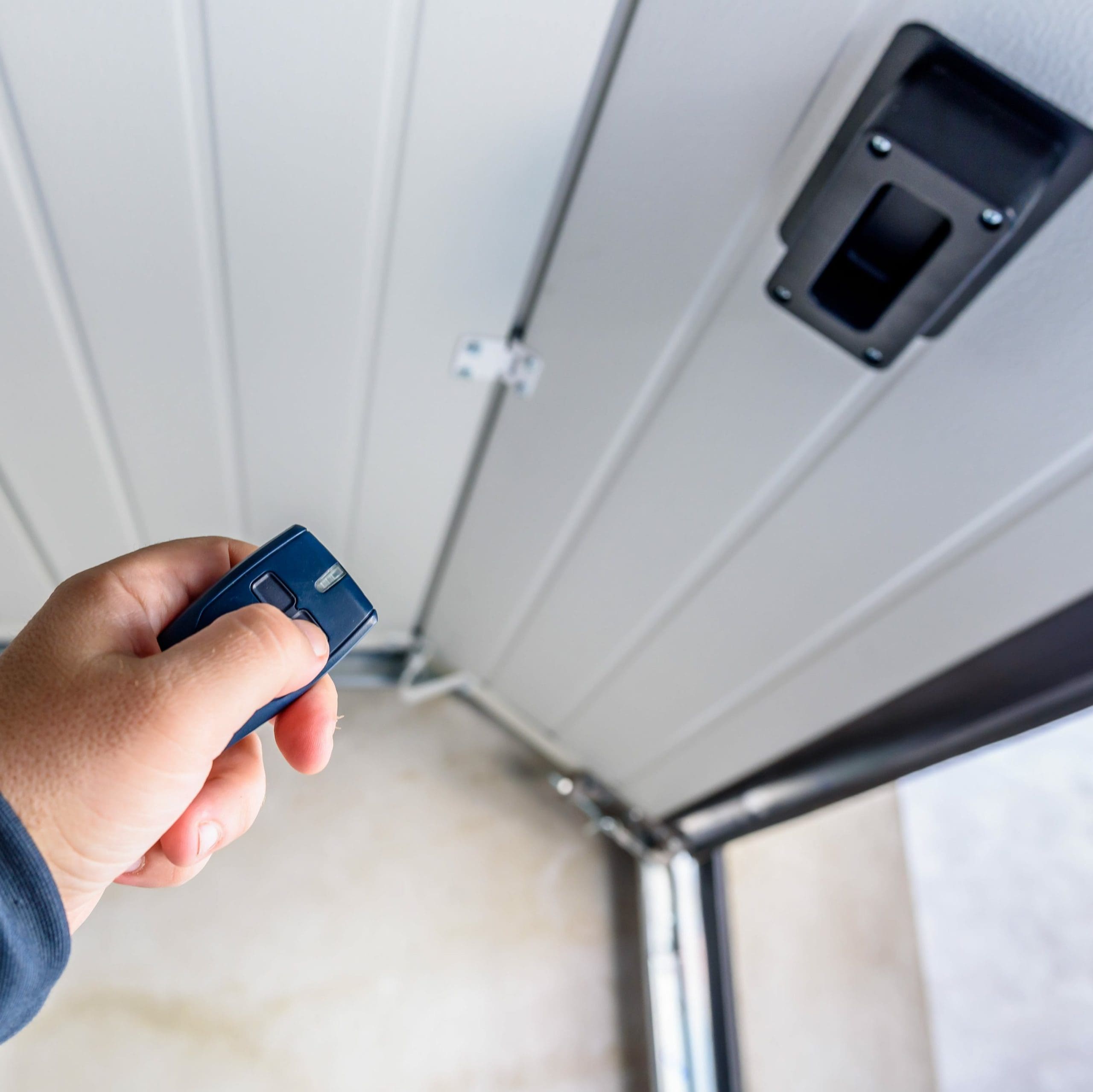 A hand holding a remote control in front of a partially opened garage door. The remote is pointed towards the door, and the door's inner wall and mechanisms are visible. The background shows the garage floor and a bit of the outdoor area.