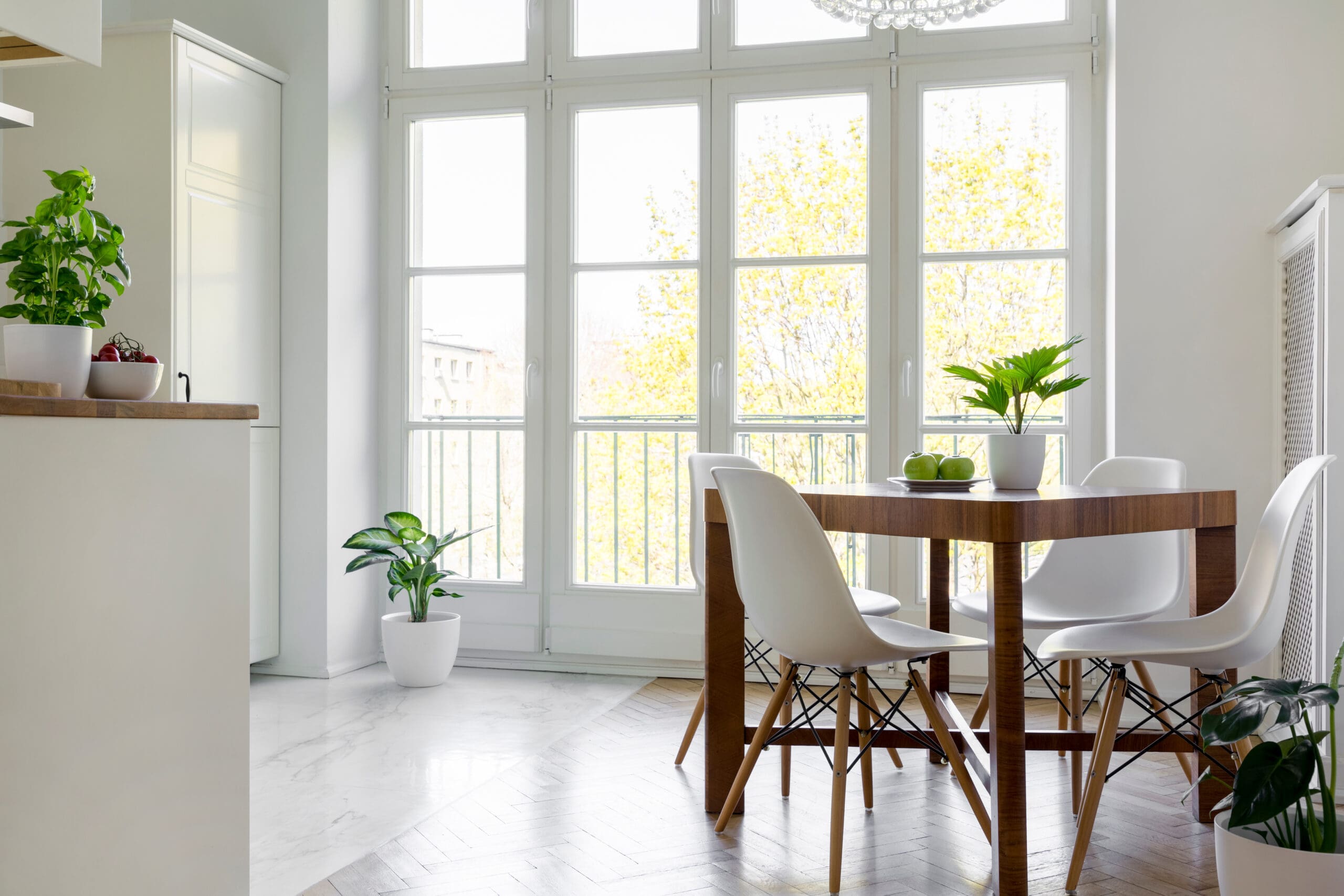 A bright, modern dining room with tall windows letting in natural light. The room features a wooden dining table with white chairs and plants adding a fresh touch. The floor is a mix of white marble and wood. A side counter with more plants is visible on the left.