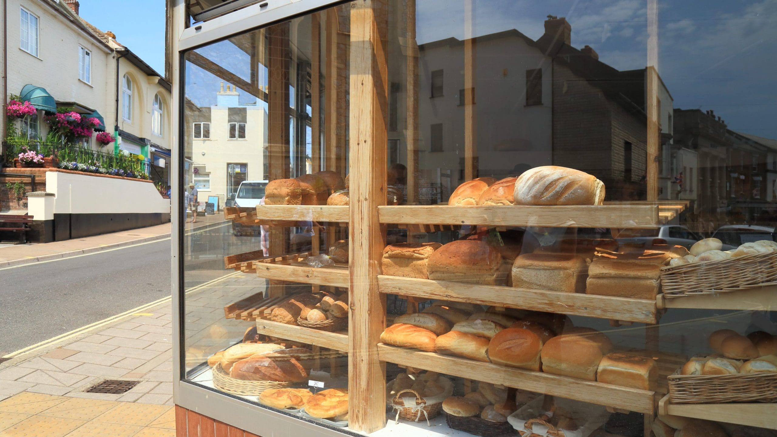 A bakery window displaying various types of bread and pastries on wooden shelves. The reflection of nearby buildings and a clear blue sky is visible in the glass. The street outside features a flower-adorned staircase and a road with cars and buildings in the background.