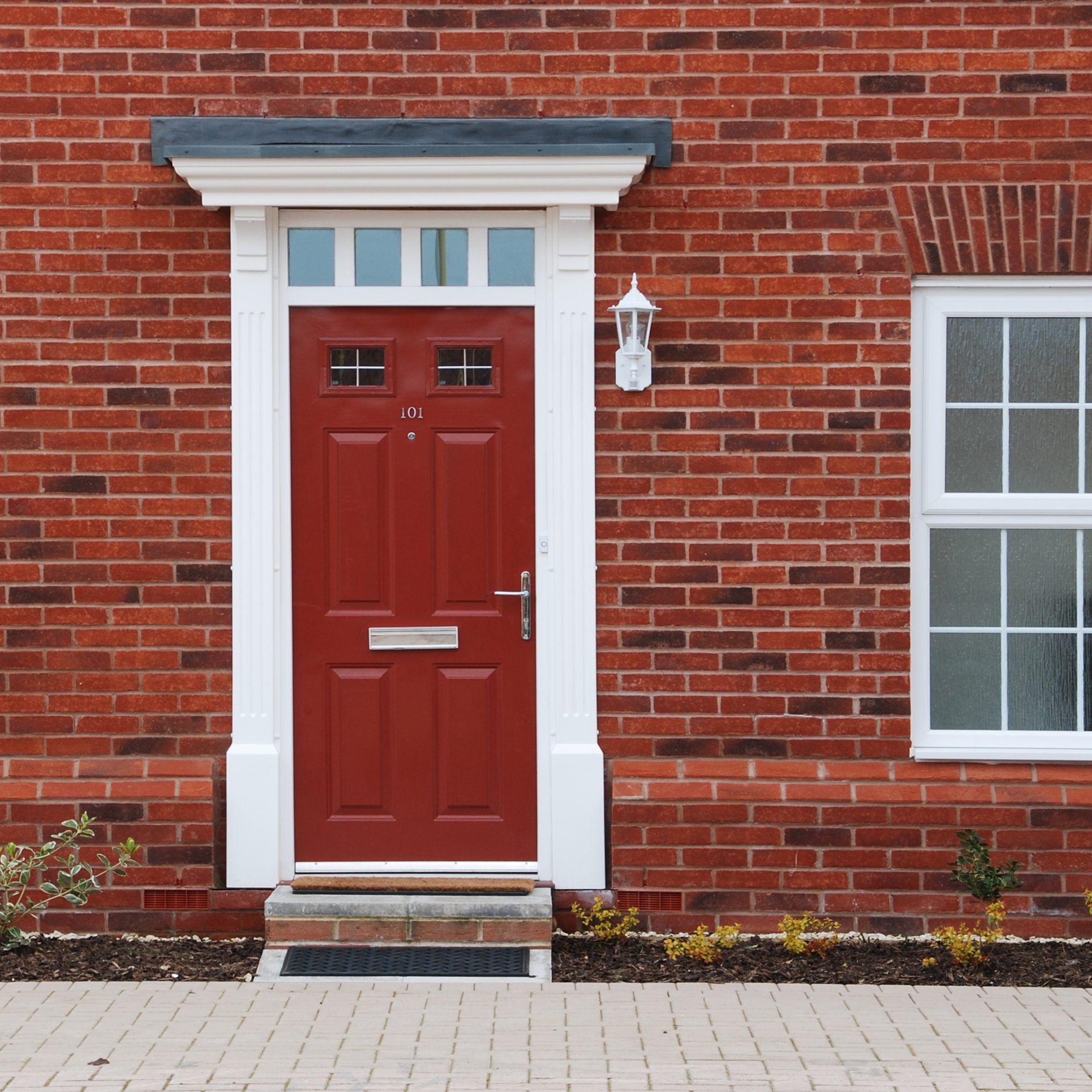 A vibrant red door with the number 101 mounted on a brick house. The door has a small mail slot and features white trim with decorative molding. To the right, there's a window with a white frame. A lantern-style light fixture is also mounted near the door.
