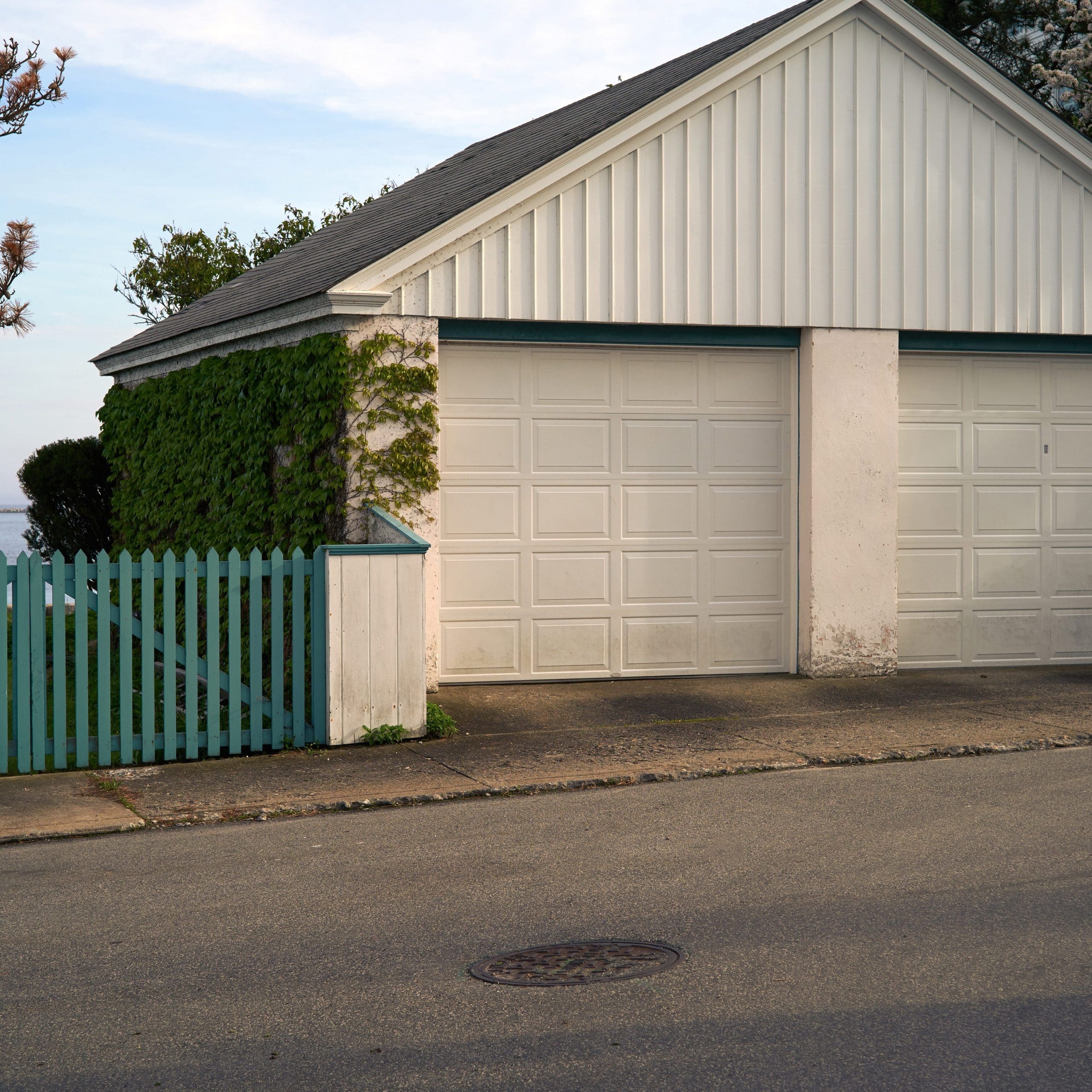 A two-car white garage with a sloped roof, adjacent to a blue picket fence on its left. Ivy grows on the garage's left side. The garage faces a paved street, with a manhole cover visible in the foreground.