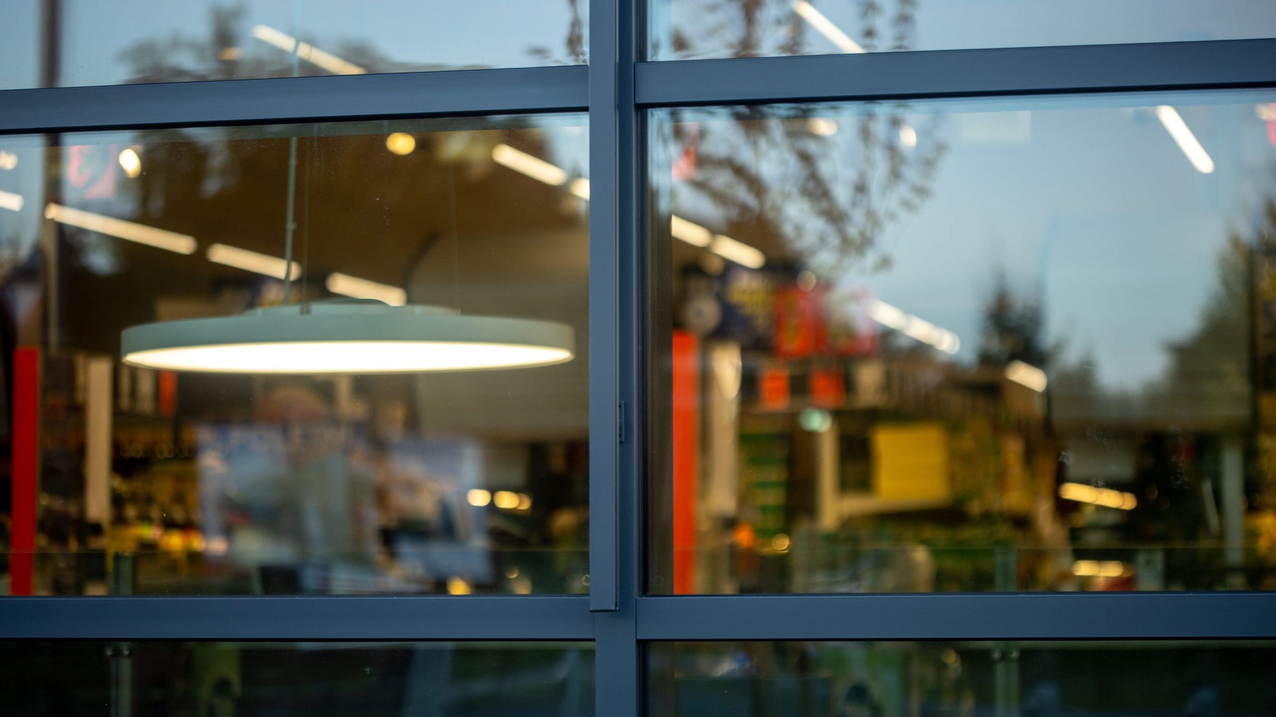 A view through large glass windows reveals the interior of a shop with bright lights, shelves, and merchandise. A hanging circular lamp is prominently visible, and reflections of trees and the sky can be seen on the glass surface.