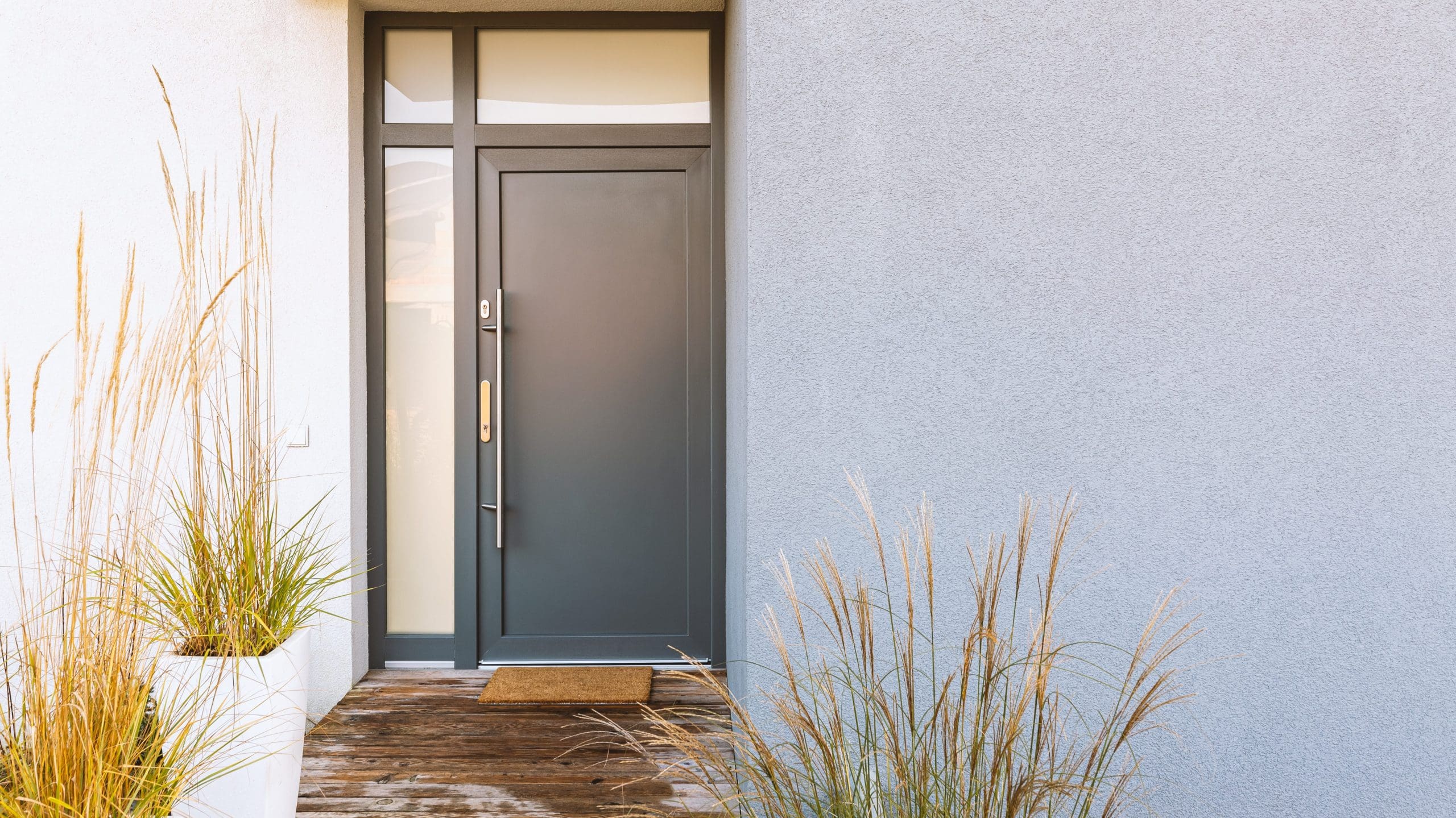 A modern gray door with a narrow vertical window beside it, set in a smooth, light gray wall. Tall grass plants in white rectangular planters flank either side of the wooden walkway leading to the door. A small doormat lies in front of the entrance.