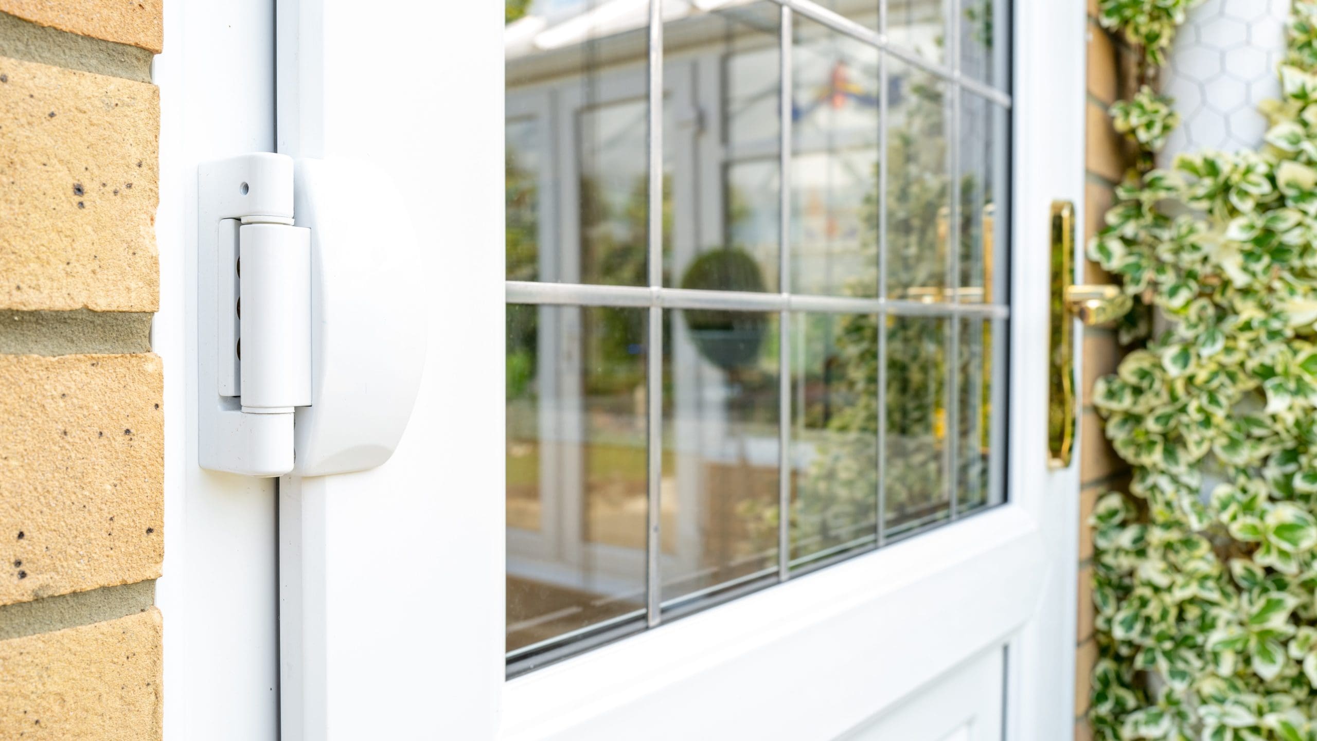 Close-up of a white door with glass panels, showcasing a metal hinge on the left. The glass reflects a green outdoor scene with foliage. The door has a gold-colored handle and is surrounded by brick and greenery. The scene appears to be bright and sunny.