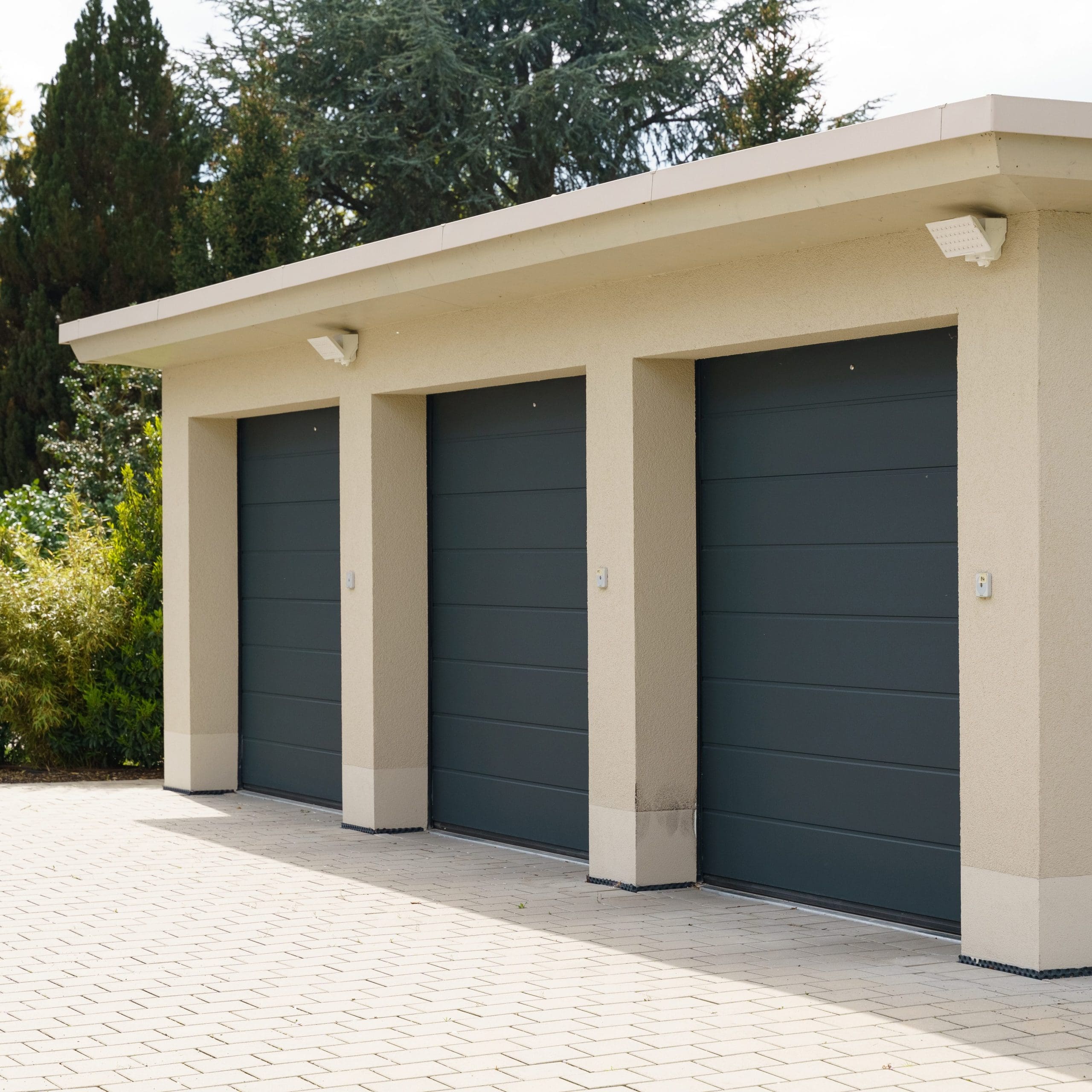 A beige building with three dark gray garage doors. The ground is paved with light-colored cobblestones, and the building is surrounded by greenery including bushes and tall trees. Each garage door has a small light fixture above it.