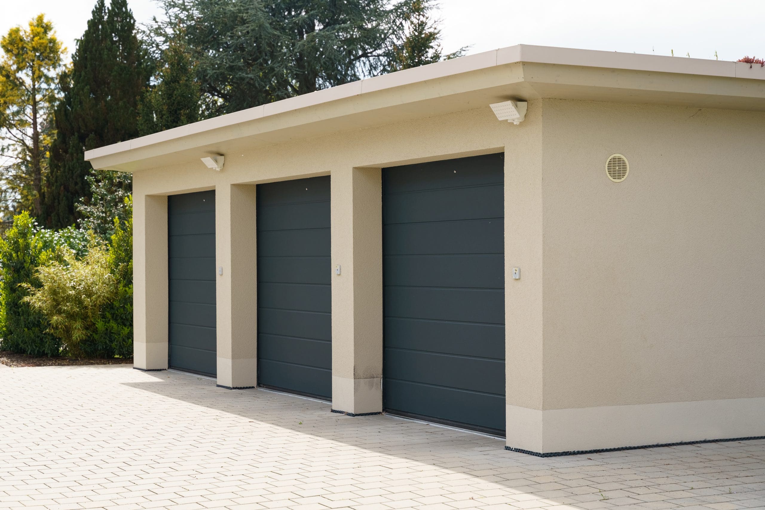 A beige building with three dark green garage doors, surrounded by greenery. The structure has a flat roof and outdoor lighting fixtures above each door. The ground in front is paved with light-colored bricks. Trees and shrubs are visible in the background.