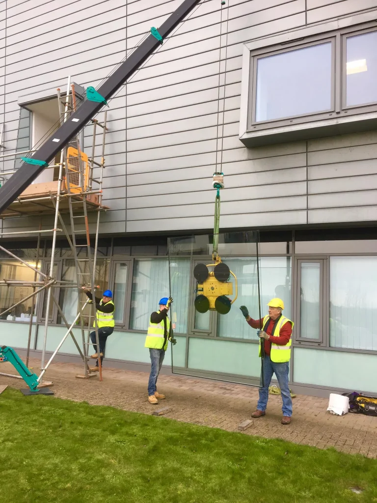 Construction workers in safety gear are installing a large glass panel on the exterior of a building. A crane is lifting the glass panel, and two workers guide it into place. Another worker stands by the crane. The building has a modern design with metal siding.