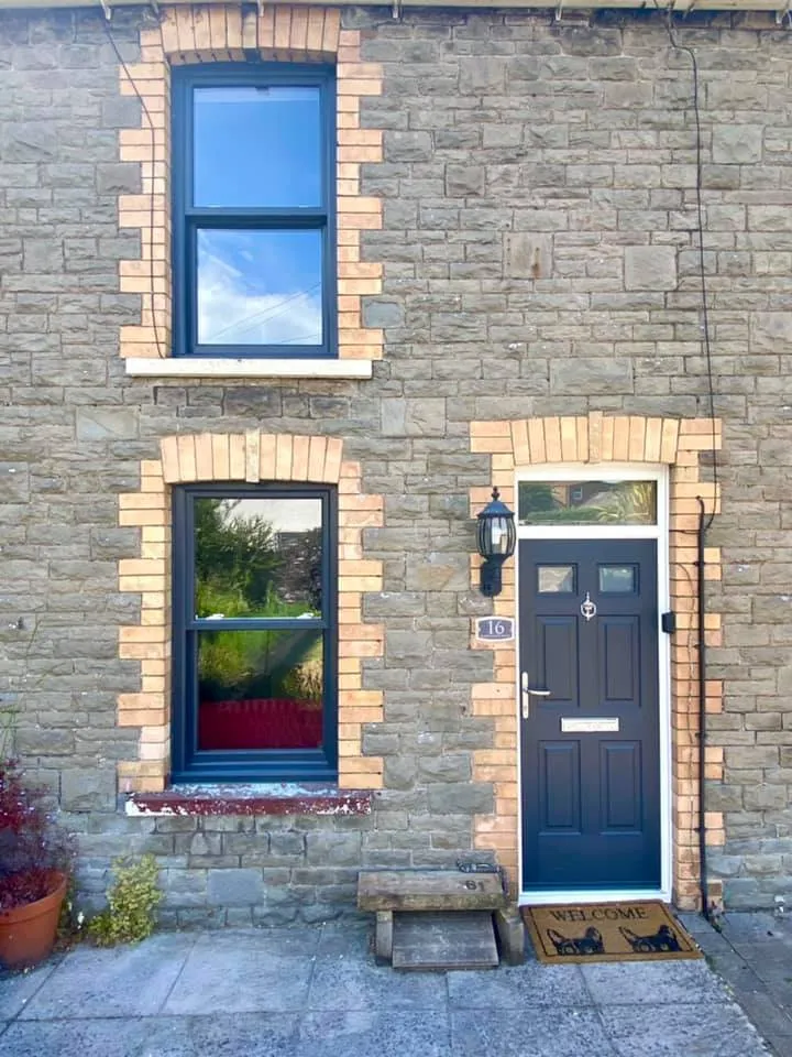 A stone house with two black-framed windows, one on the ground floor and one directly above it. The house has a dark blue door with a lantern on the right side, a doorbell, a house number plaque with "16", and a "Welcome" doormat with cat silhouettes. A small wooden step is in front of the door.