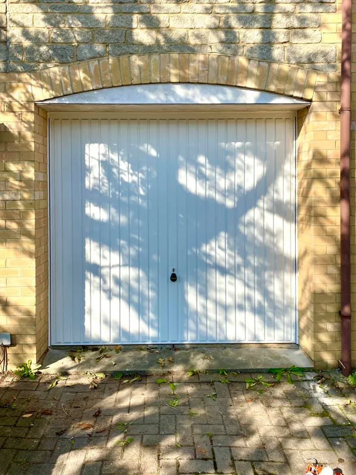 A closed white garage door set in a brick wall, partially shaded by tree branches and leaves. There is a small circular handle in the middle of the door, and scattered leaves are on the paved ground in front. The shadow of a tree is cast prominently on the door.