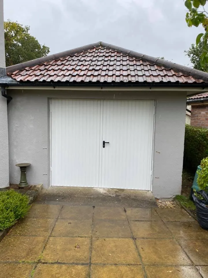 A single-car garage with a peaked, tiled roof and white, vertically paneled doors, surrounded by bushes on either side. There is a small birdbath on the left. The ground in front is paved with wet square tiles, indicating recent rain.