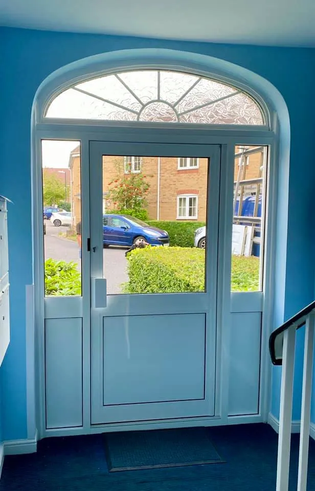 A view from inside a blue-walled foyer looking out through a white-framed glass door with an arched window at the top. Outside, there's a blue car parked in front of a brick building, and greenery is visible. A staircase is partially visible on the right.