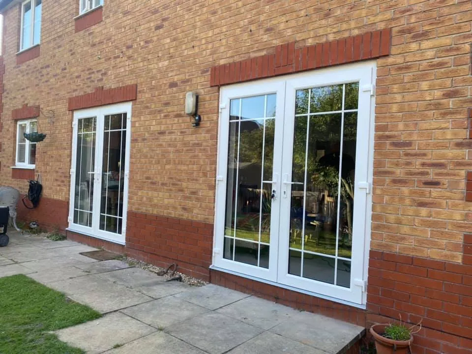 A brick house exterior featuring three white-framed glass doors. Two are paired French doors, and the third is a single door with a decorative grid pattern. There is a small grassy area, a potted plant, and a patio with stone tiles in front of the doors.
