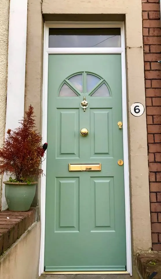 A light green front door with a semi-circular window at the top, brass door knocker, mail slot, and door handle. The door is framed with light-colored trim, and there's a potted plant to the left and the number 6 plaque to the right on a brick wall.