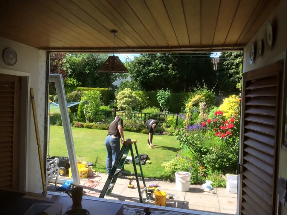 Two people are working in a garden viewed through an open window from inside a house. One person is using a ladder and tools, while the other is bending over, tending to the plants. The garden is lush and green with blooming flowers and bushes.