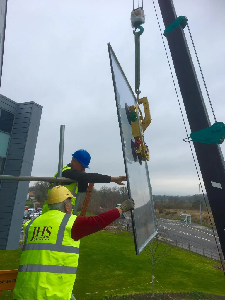 Two construction workers in high-visibility vests are installing a large glass pane using a crane on a multi-story building. One worker is on a ladder, guiding the glass, while the other holds the pane. An expansive view of greenery and a road is seen in the background.