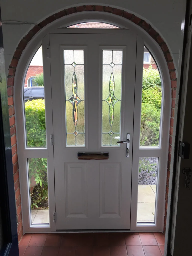 A white front door with decorative vertical glass panels and a mail slot stands framed by an arched brick entryway. Windows on either side and above the door let in light. Through the glass, greenery and part of a neighboring house are visible outside.