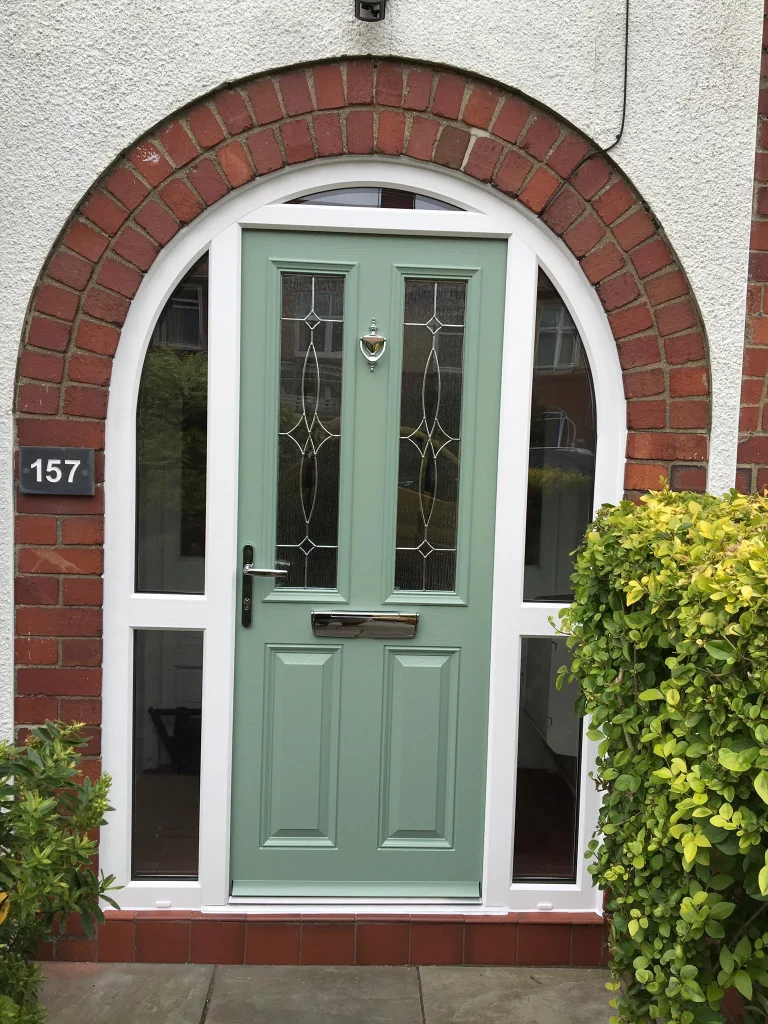 A green front door framed by a red brick archway with white trim. The door has two narrow, decorative glass panels, a mail slot, and a silver door knocker. The house number 157 is displayed on a plaque to the left. Green bushes are on the right side.