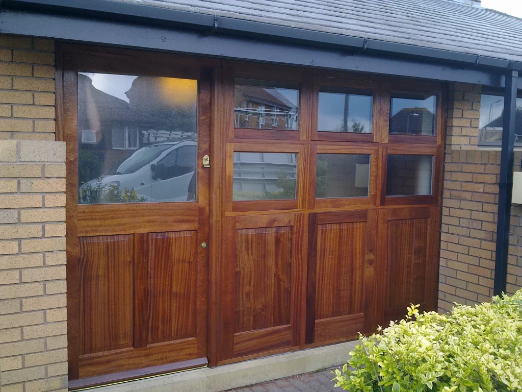A sleek wooden garage door with multiple glass panels on the upper half is set into a brick exterior. The door reflects sunlight, highlighting its polished finish. A parked white van is slightly visible through the glass panels. Green shrubs line the area in front.