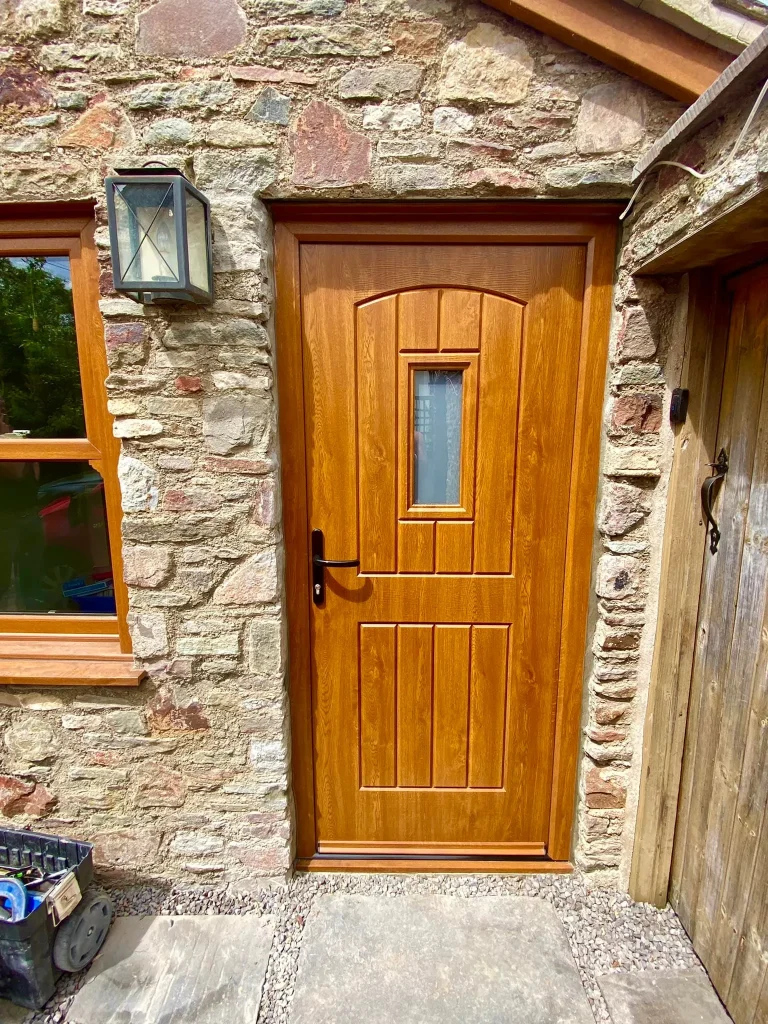 A wooden front door set in a stone wall with a rectangular window in the upper part. To the left, there's a lantern-style light fixture and a wooden-framed window. The door has a handle and lock, and there are stone steps leading up to it.