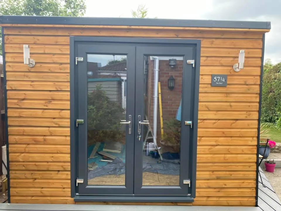 A wooden shed with large glass double doors is seen. The shed has horizontal wooden siding and a flat roof. Two outdoor lights are mounted on either side of the doors, and the number 571 is visible to the right of the doors. Plants are seen to the right.