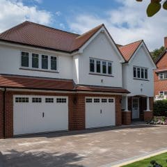 A two-story suburban house with a white facade and red brick accents features a three-car garage with white doors. The house is surrounded by neatly landscaped greenery and a paved driveway under a partly cloudy sky. Another house is visible to the right.
